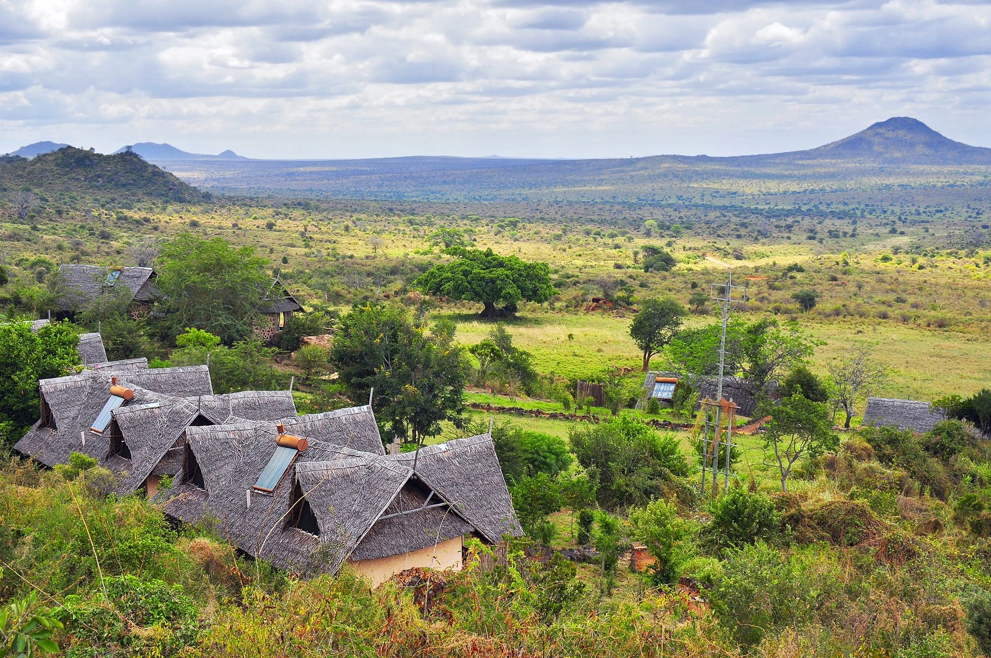 Thatched lodge cottages on a hillside above green savanna and volcanic hills